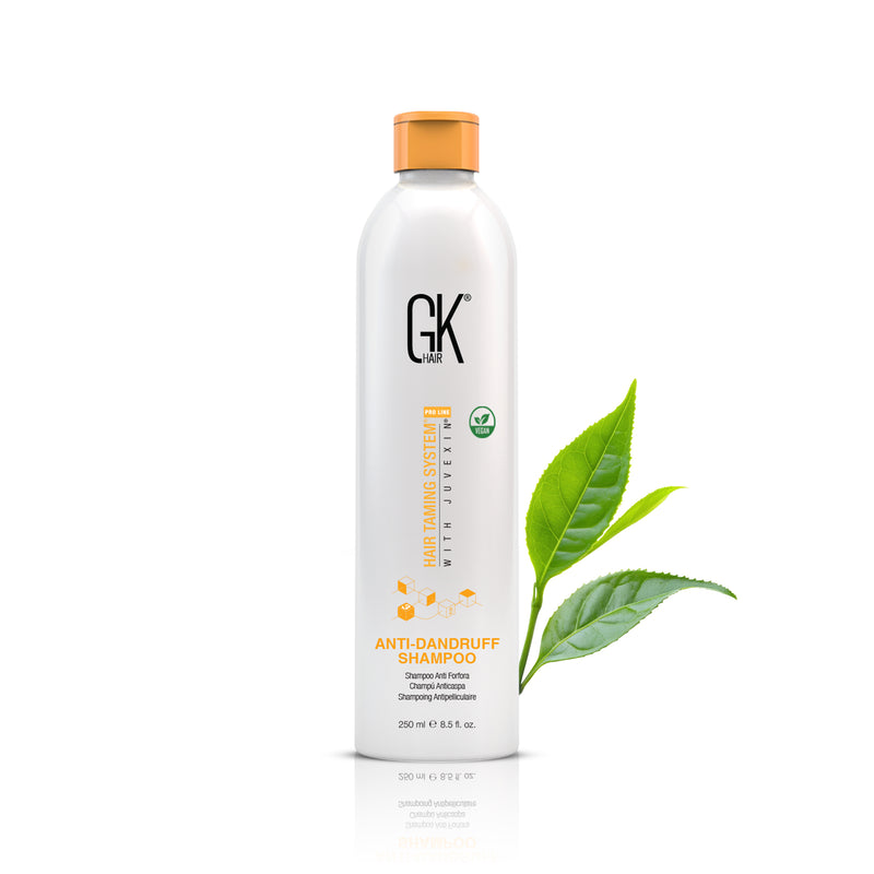 GK Hair Anti-Dandruff Shampoo 250 ml white bottle with an orange cap, front view beside fresh green tea leaves