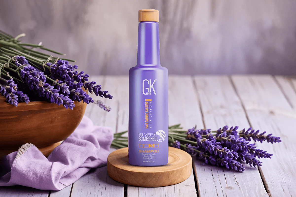 Purple shampoo bottle labeled "GK Hair Silver Bombshell" on a wooden stand, surrounded by lavender flowers and a wooden bowl, on a rustic table.